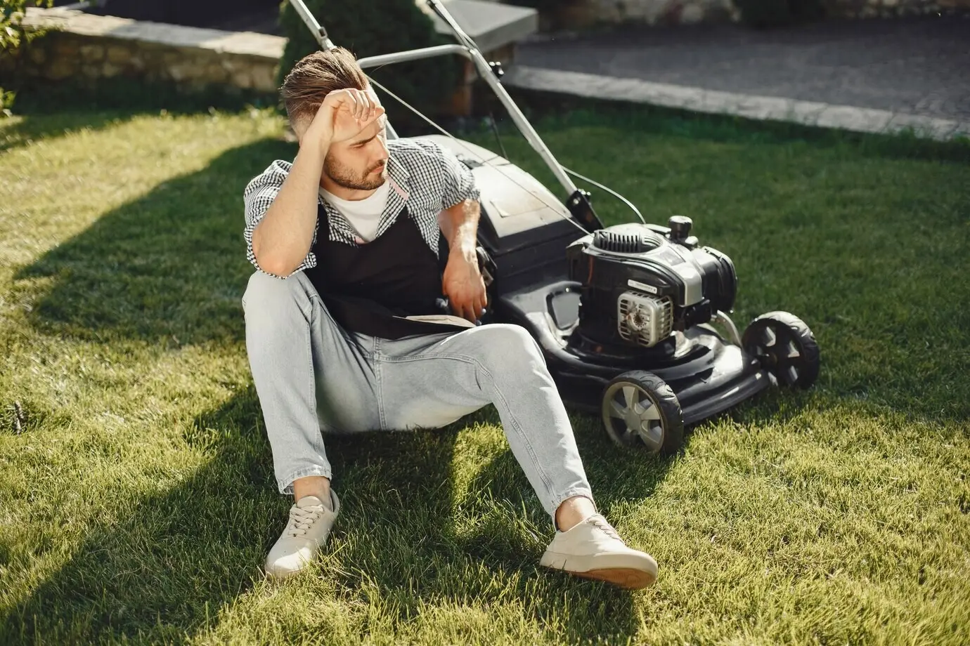 A man wearing a black apron is mowing the grass in the backyard with a lawn mower.