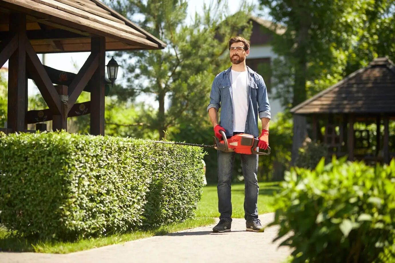 Low-angle view of a happy bearded gardener standing in a park, carrying a modern handheld hedge trimmer.